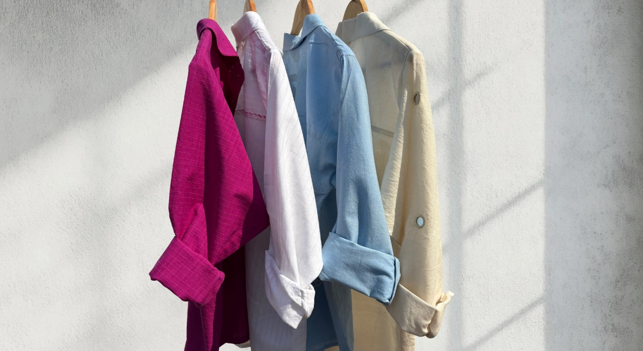 Four shirts in pink, white, blue, and beige hanging on a rack against a light gray wall.