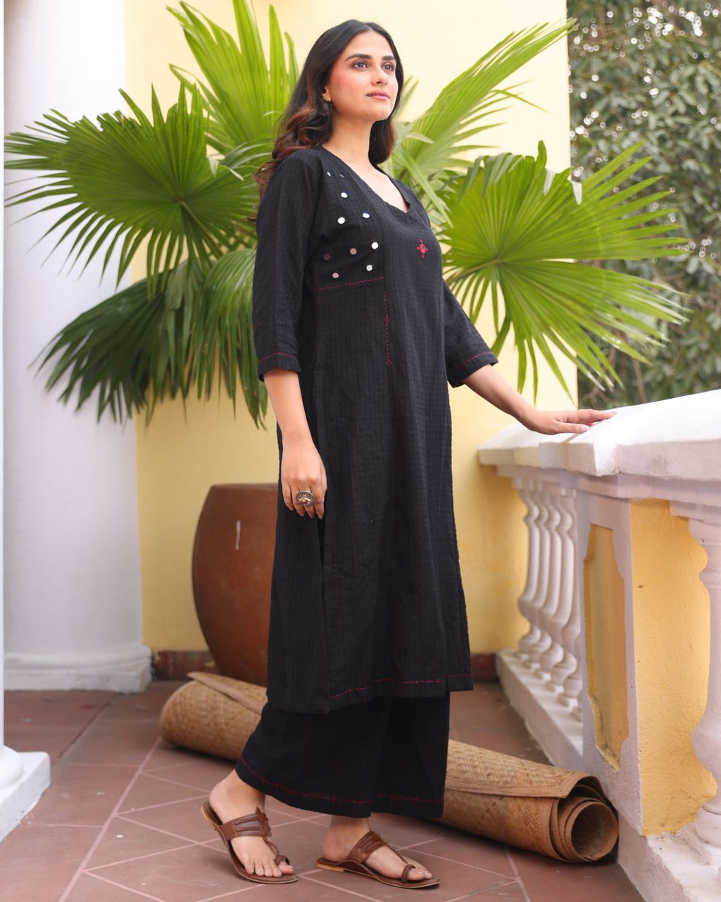 Woman in a black outfit standing in front of green plants and a yellow wall.