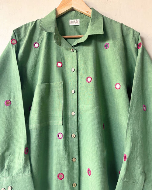 Green shirt with pink button details on a hanger against a beige wall.