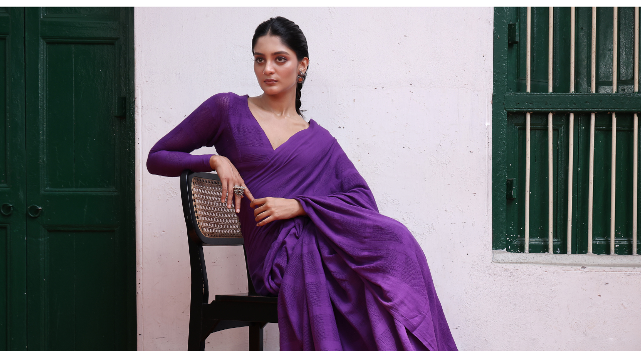 Woman in a purple saree sitting on a chair against a white wall with green doors.