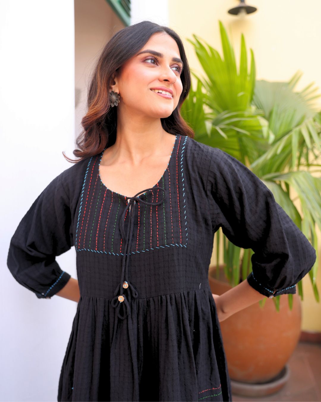Woman wearing a black dress with decorative trim in an outdoor setting with plants.